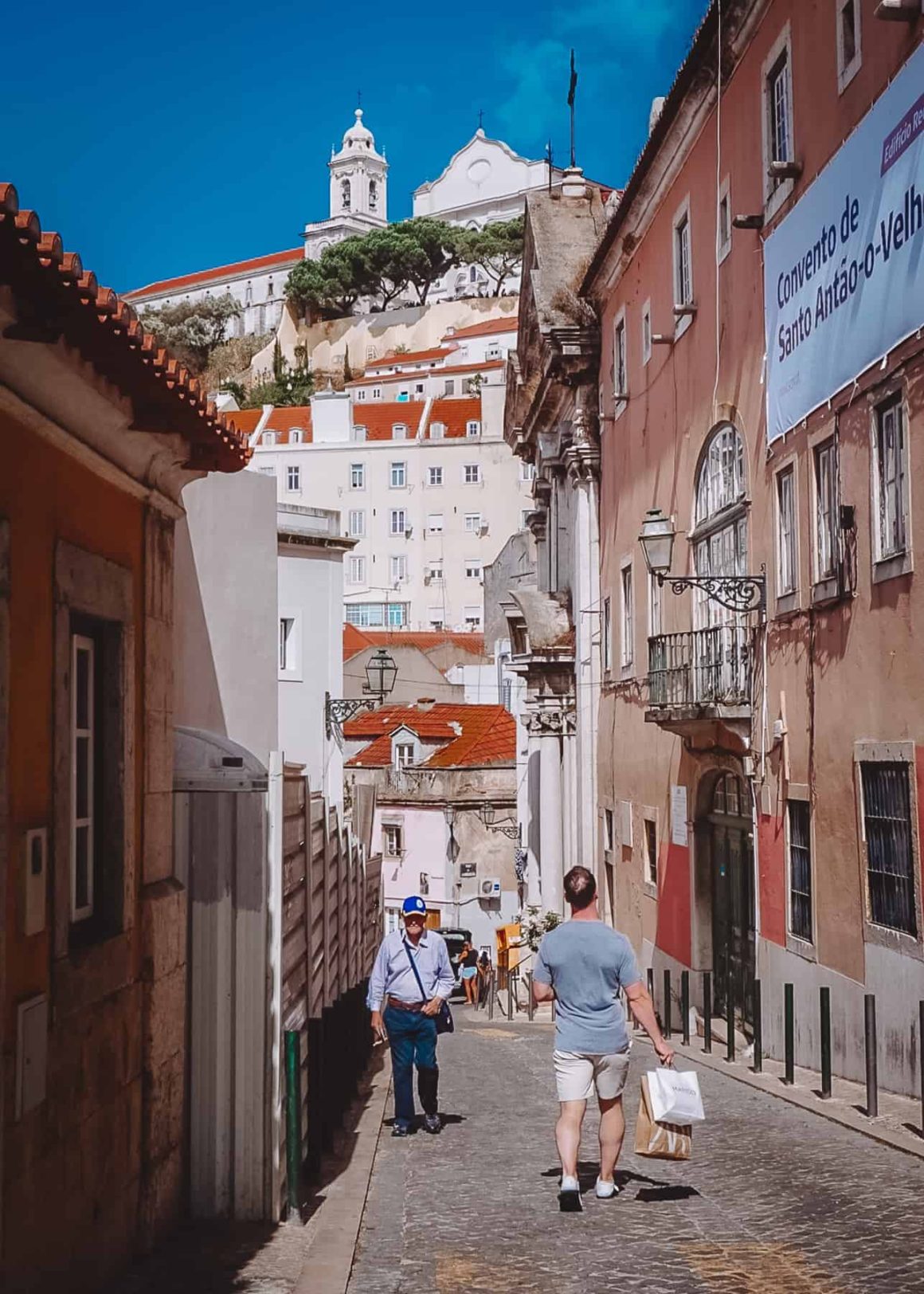 A cobblestone street with people walking surrounded by houses and buildings up until the hill in the background under blue skies