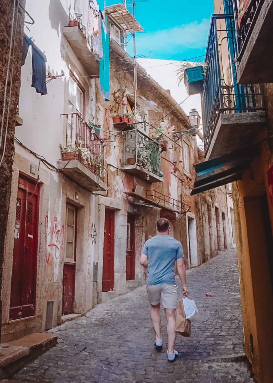 A narrow street where a person is passing by surrounded by old buildings with red colored doors and small balconies, a common sight to see in your 2 days in Lisbon stay