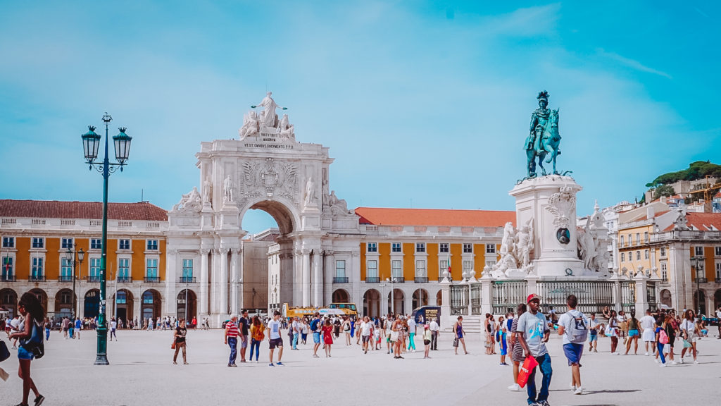 The city square in the Lisbon with people walking while surrounded with a statue and colorful buildings under blue skies