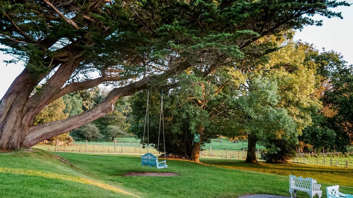 A peaceful garden swing beneath mature trees overlooking the landscaped grounds and countryside.
