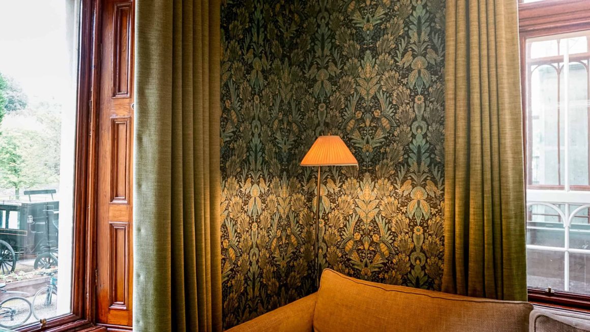 Cosy lounge corner with patterned wallpaper, tall windows, green curtains and a floor lamp inside a historic country house.