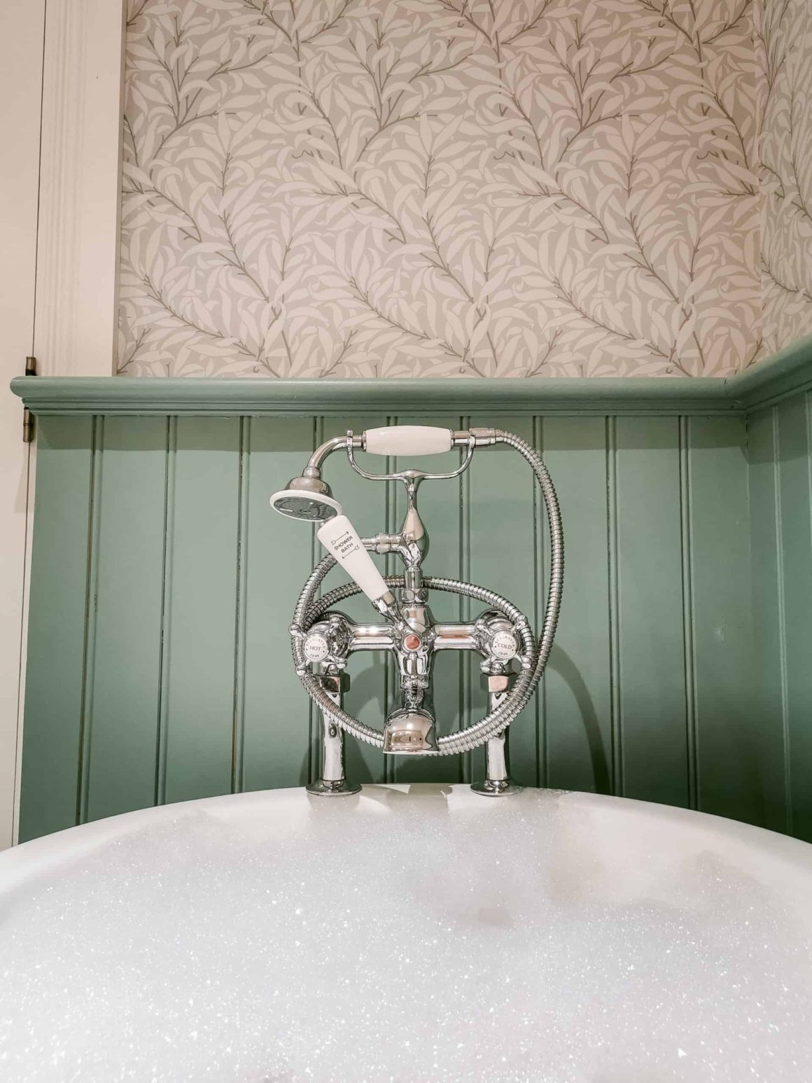 Traditional roll-top bath filled with bubbles and vintage brass fittings in a beautifully designed bathroom.