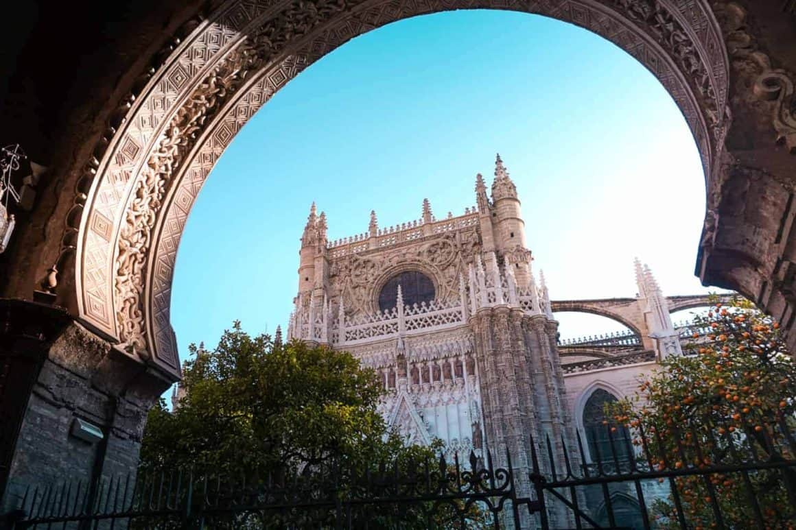 Ornate Gothic exterior of Seville Cathedral framed by an archway and orange trees beneath a clear blue sky.