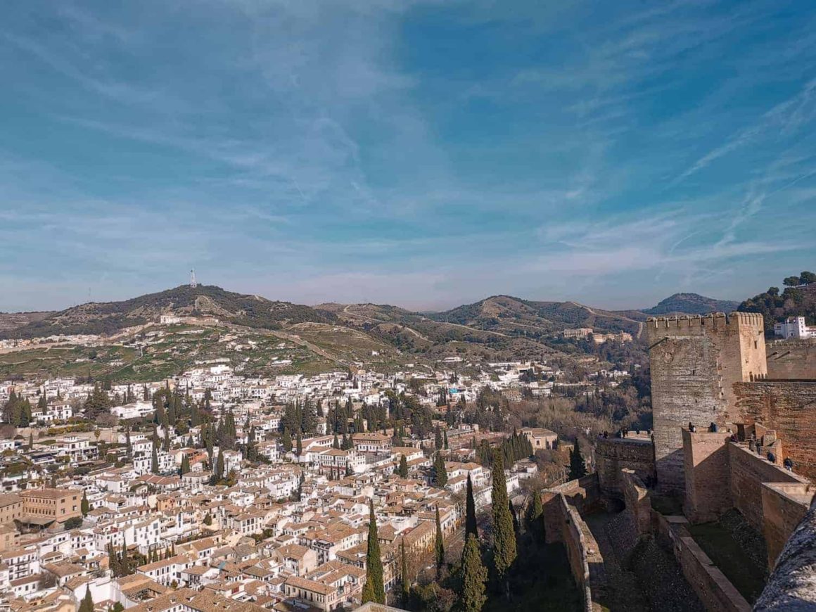 Panoramic view across Granada with whitewashed buildings, rolling hills and the historic Alhambra fortress walls.
