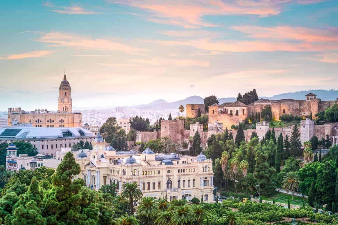 Panoramic view of Mรกlaga with the historic Alcazaba, cathedral tower and city buildings beneath a colourful sunset sky.