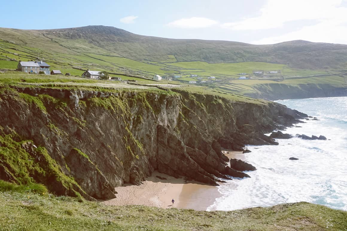 Rugged sea cliffs surrounding a small sandy beach, with green fields, scattered houses and rolling hills along the Irish coast.