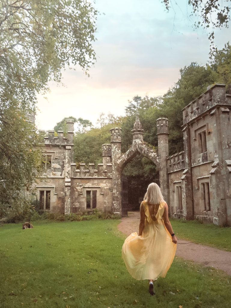 Woman in a yellow dress walking towards weathered Gothic castle ruins surrounded by trees and green lawns in Ireland.