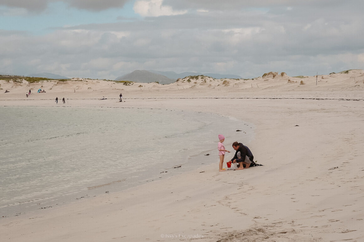 An adult kneeling beside a small child on a wide sandy beach, with dunes and distant mountains under cloud