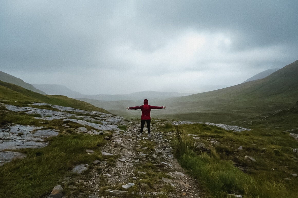A person in a red jacket standing with arms outstretched on a rocky trail overlooking a misty mountain valley