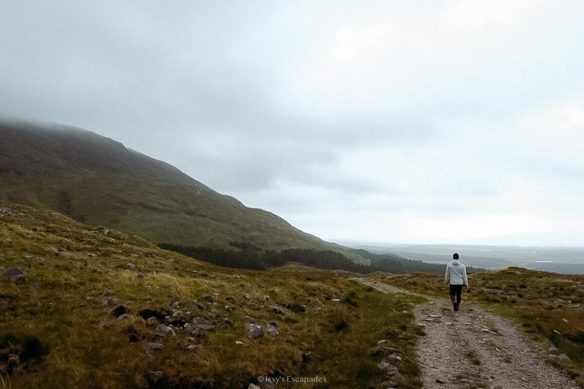 A person in a light jacket walking along a gravel mountain track beneath low grey cloud