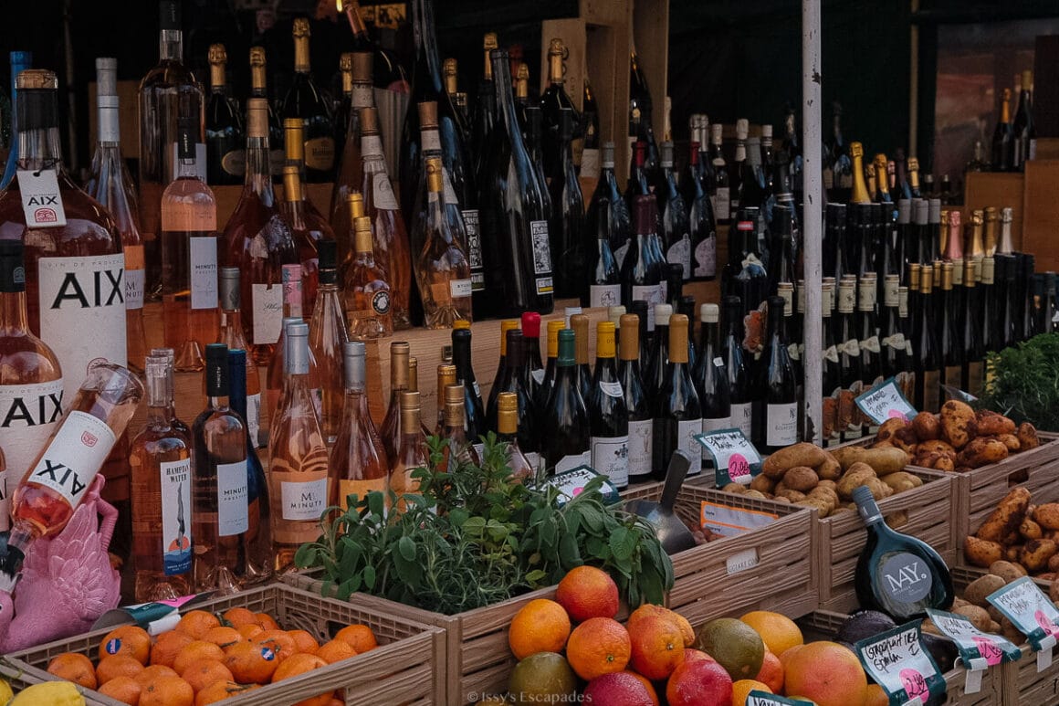Market stall in Munich displaying bottles of wine alongside fresh fruit, vegetables and other produce.