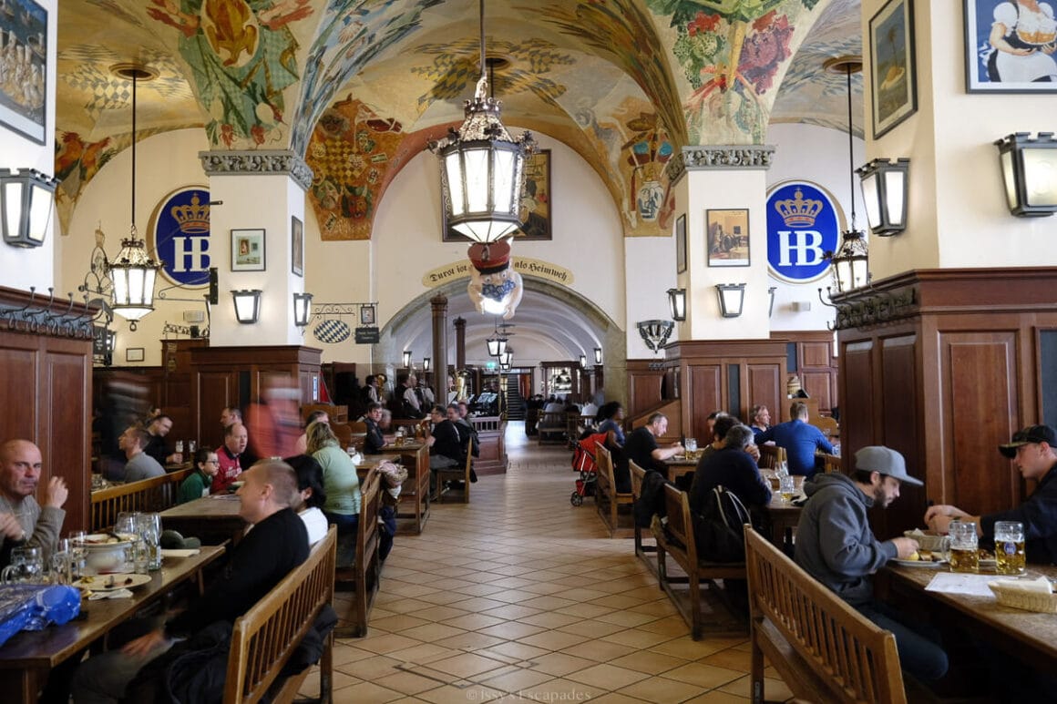 Traditional interior of the Hofbrรคuhaus beer hall in Munich with long wooden tables, diners and a decorated vaulted ceiling.