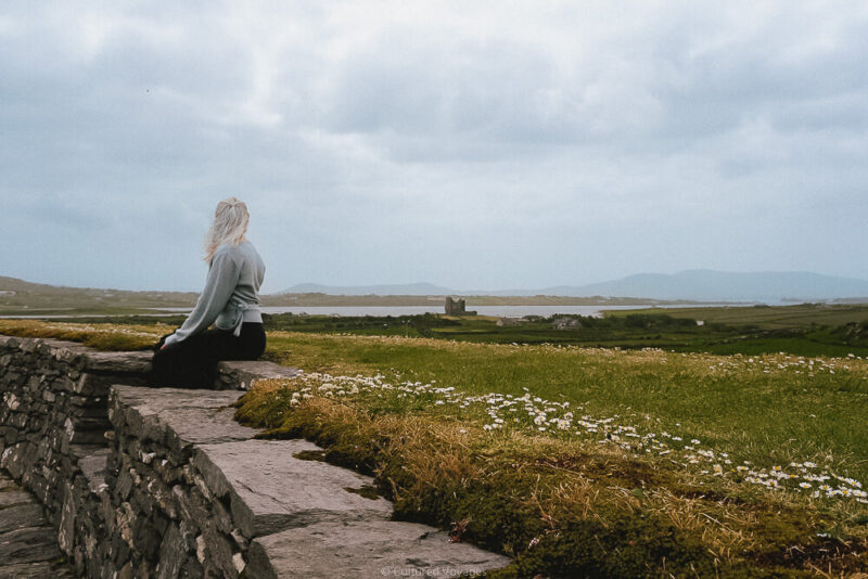 A woman sits on the wall of Cahergal Fort looking towards Ballycarbery Castle