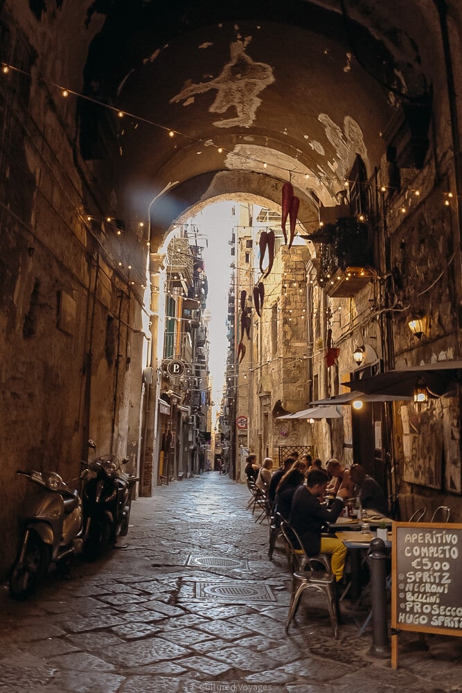 archway leading down narrow alley in naples with people sitting having drinks on one side