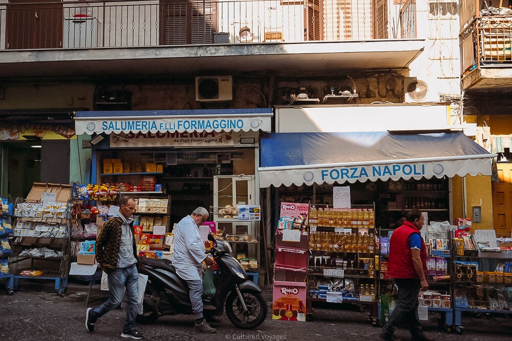 men walk in front of colourful streetside food stall in naples