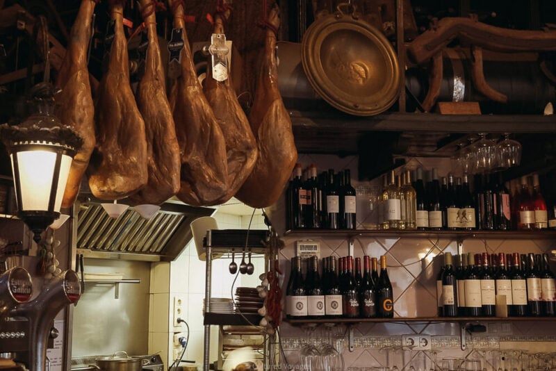 Cured hams hang above a bar filled with bottles and glasses