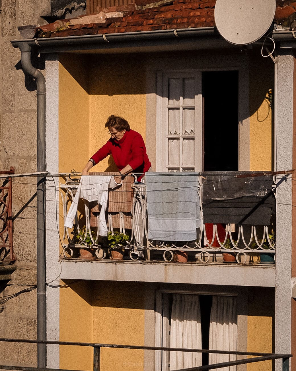 A person hanging mattresses and clothes on a balcony of a yellow with red tiled roof house