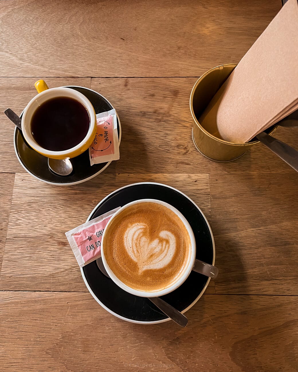two cups of coffee served with teaspoons, sugar packets, and tissues on top of a wooden table in Nicolau Porto