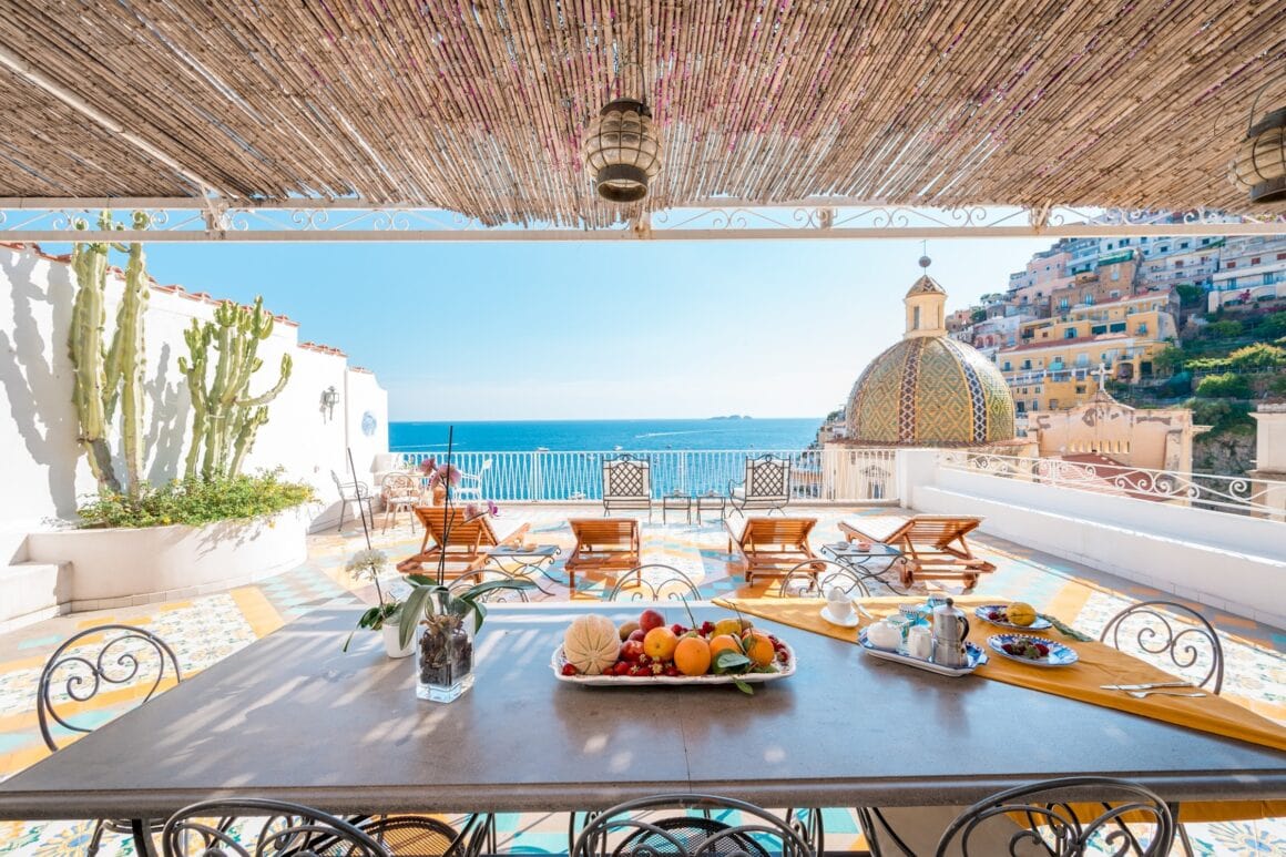 A sunlit terrace overlooking the Amalfi Coast, with colourful tiled floors, sun loungers, a breakfast table and a patterned church dome in Positano, featured among things to do in ravello italy.