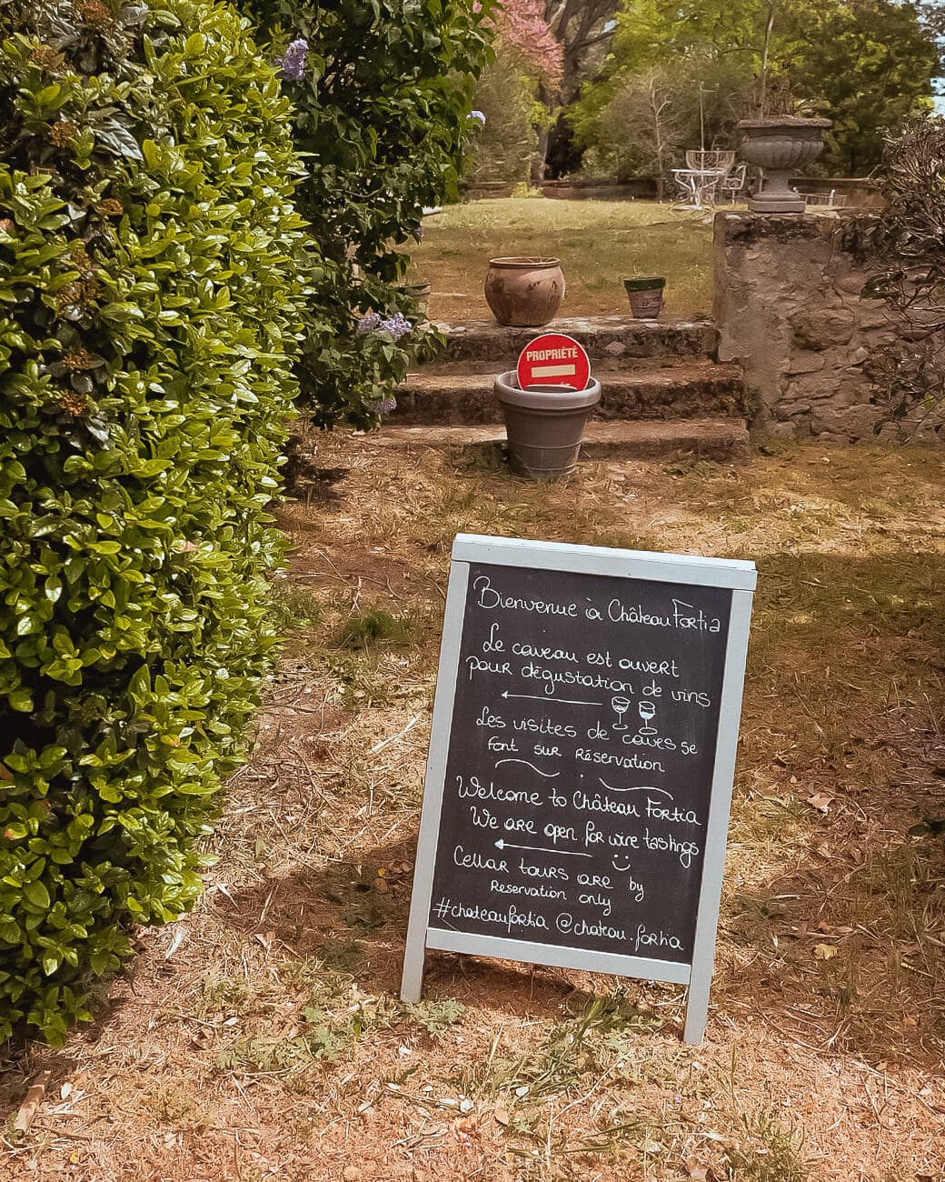 A chalkboard on a dirt road with pots in the back leading to an outdoor space in the background surrounded by trees