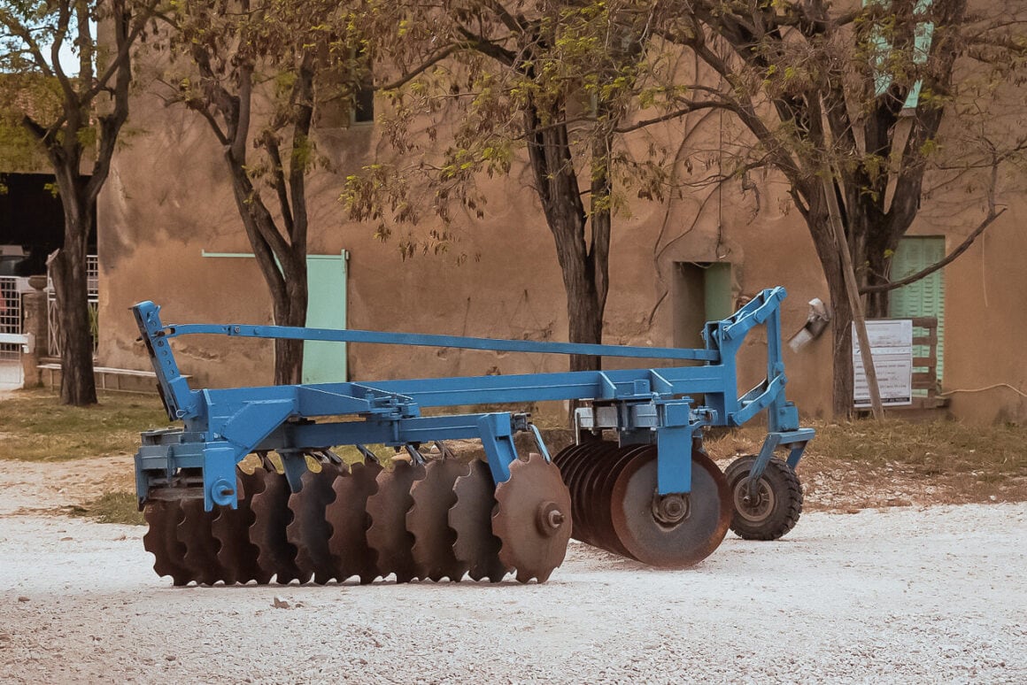A blue trailed disc harrow in blue in the center of a dirt road with trees and a brown walled building with windows and doors