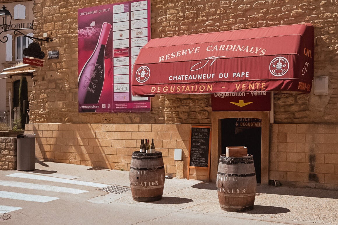 A stone bricked wall of a building beside a paved road with two wine barrels nearby the entrance that has a maroon awning with Châteauneuf-du-Pape in a white text and a wine poster on the wall