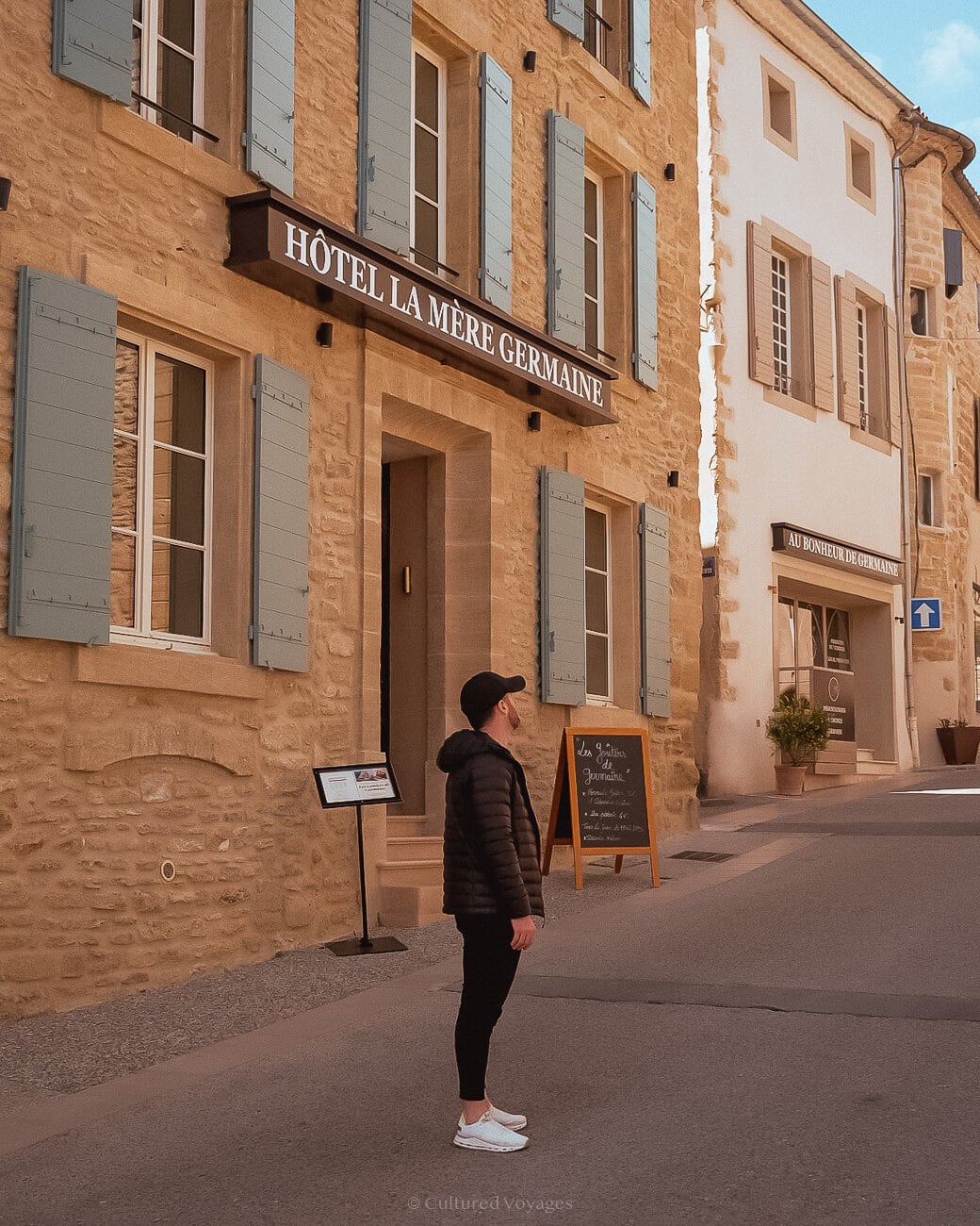 A person in a black padded jacket, black pants and hat, and white pair of shows in the middle of a paved road with a stone bricked wall hotel in the background with a chalkboard nearby the doorway
