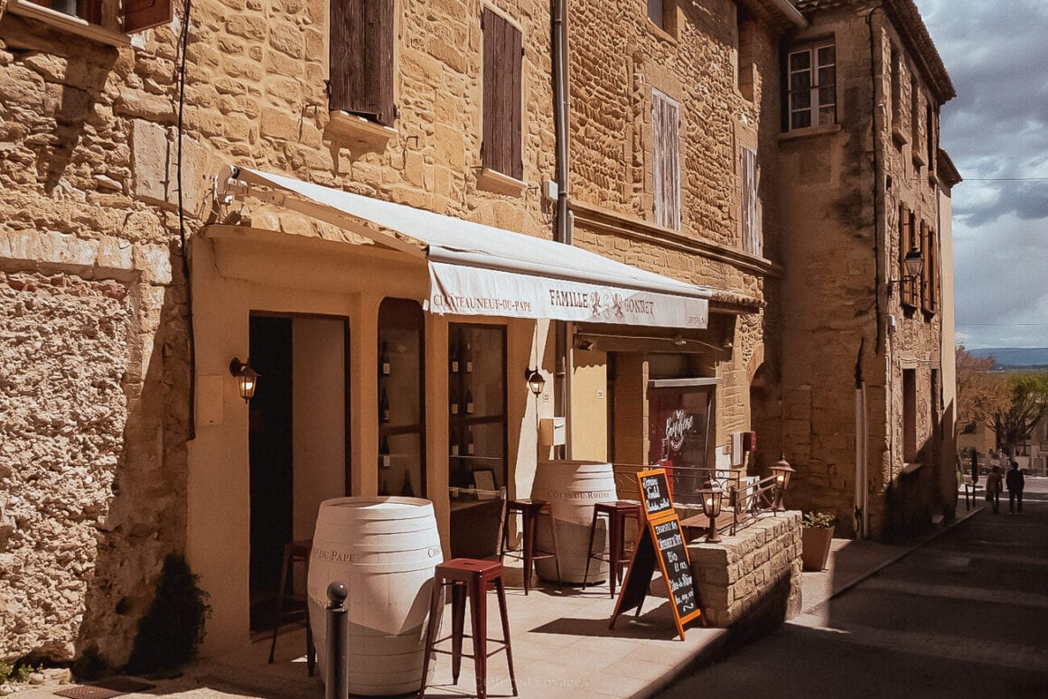 A view of a restaurant outdoor space with white painted wine barrels as tables, brown chairs, and chalkboard nearby a paved road