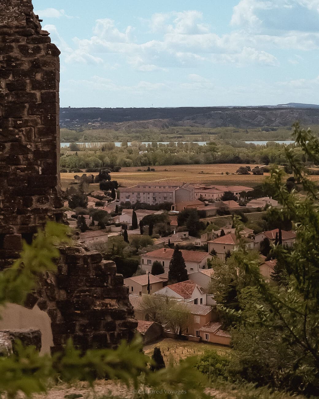 A view of a town with white houses and red-tiled roofs through bushes and ruins. In the background are a line of trees and bright skies with clouds