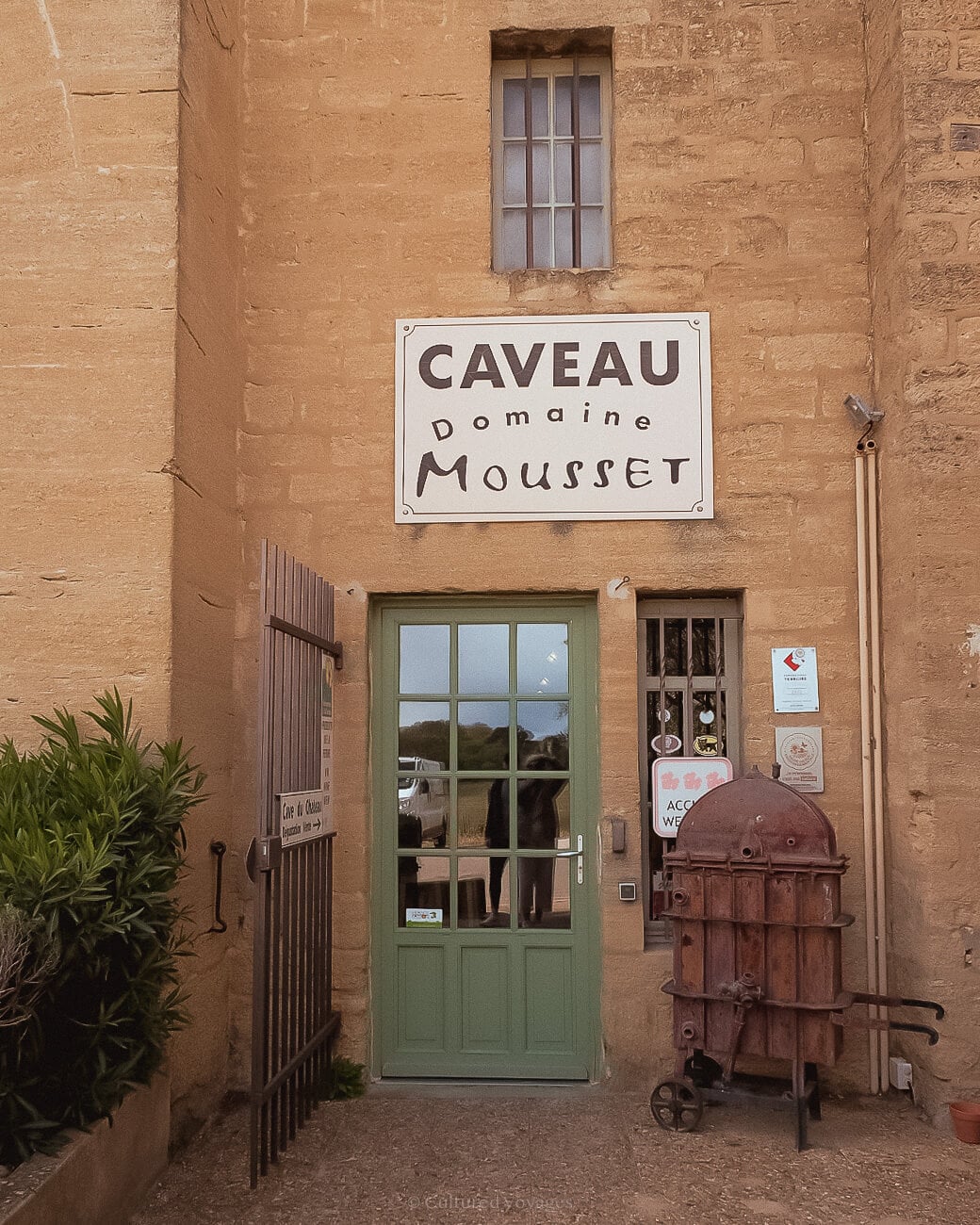 A green door with glass panels and an open steel gate, surrounded by a stone-brick wall with rectangular windows. A white sign reading "CAVEAU Domaine Mousset" hangs above the doorway.
