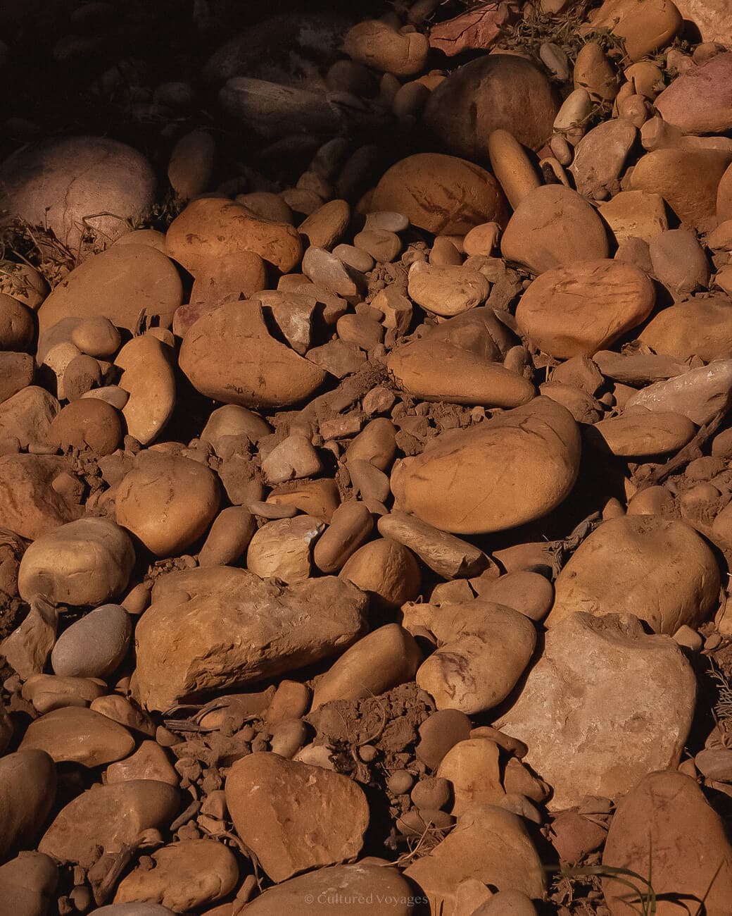 A portrait view of pebbles and stones of various sizes, covered in dry mud and a shadow on some of them