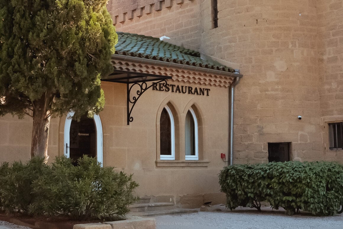 The exterior of a restaurant with a Gothic-arched doorway and windows, surrounded by hedges and a tree. The building is connected to a stone-bricked structure on one side.