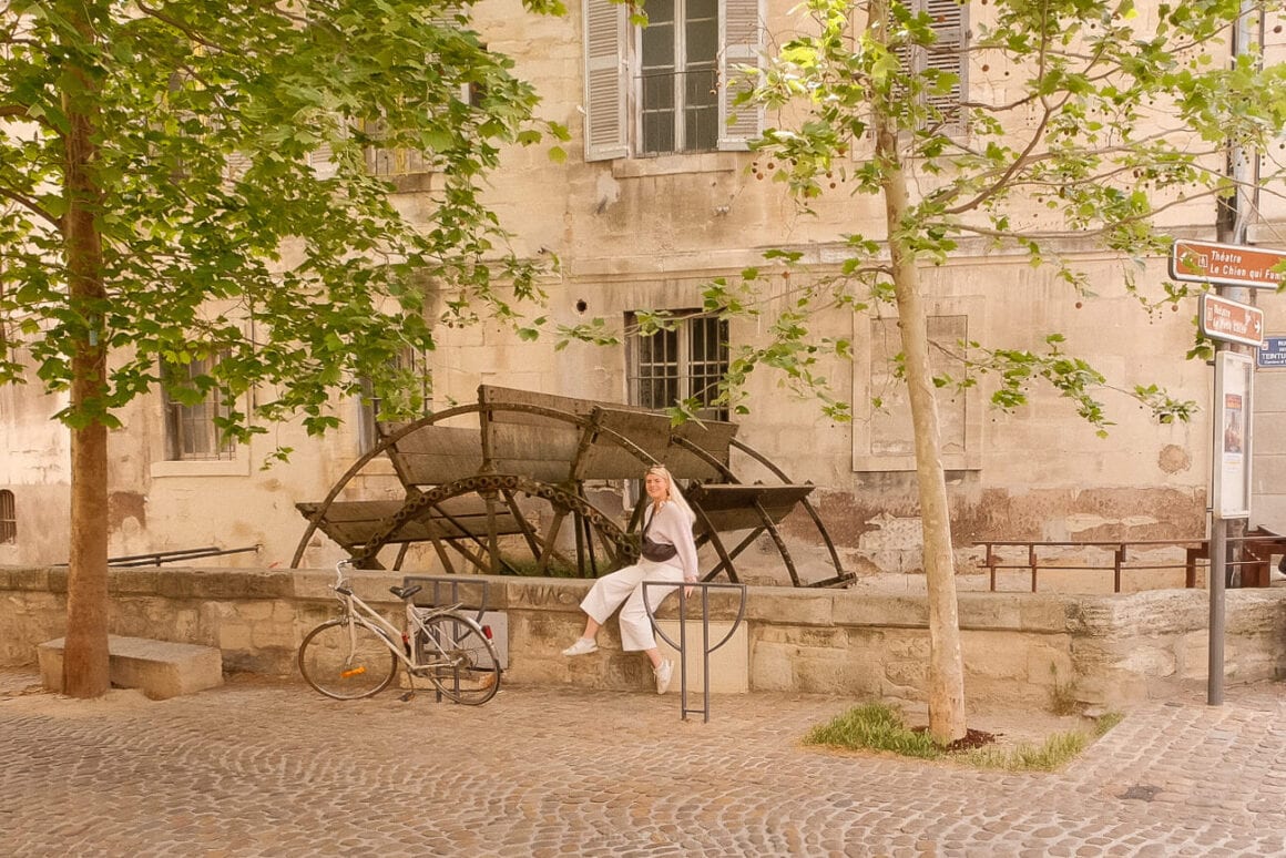 Me sitting on a low wall with a parked bicycle and trees nearby. In the background, a water wheel and building facade with windows stand. Cobblestone street lies in the foreground.