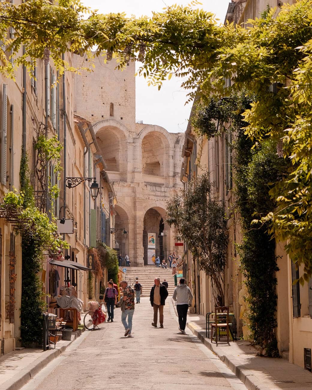 Pedestrians walking on a paved street. A vine spans across buildings, creating an entrance. In the background, a staircase leads to arched entrances of a domed structure.