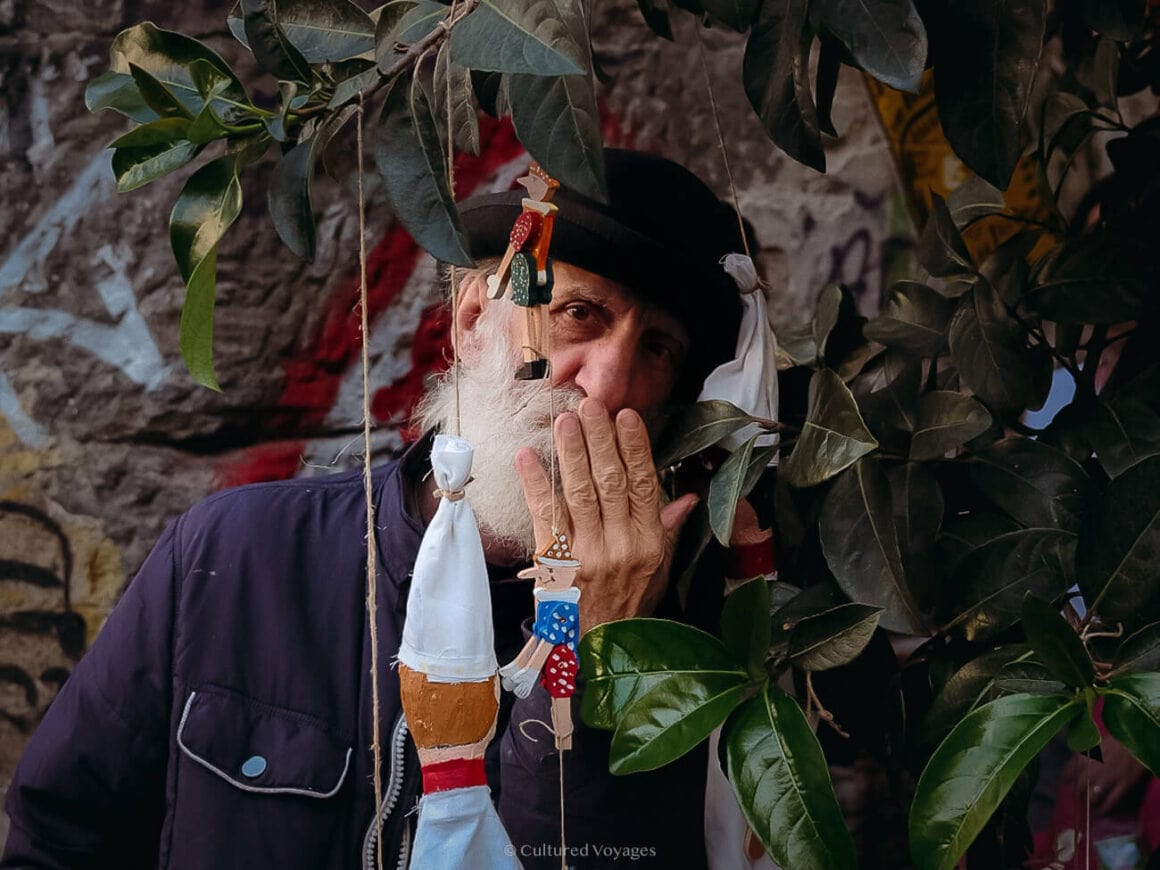 A man in a hat and blue longsleeve the surrounded by leaves and small hanging wooden dolls