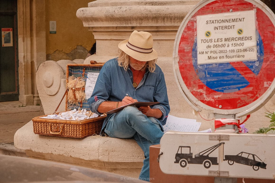 A man in jeans, blue long sleeve shirt, and straw hat sits beside a wicker suitcase filled with lavender items. In the foreground, a traffic sign displays towing instructions and other guidelines.