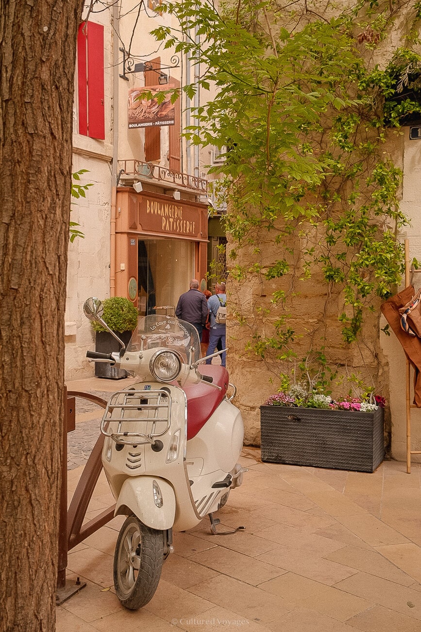 Parked white Vespa beside a tree trunk outdoors. Vines creep along a stone-bricked wall, while a white building with red window shutters appears in the background.