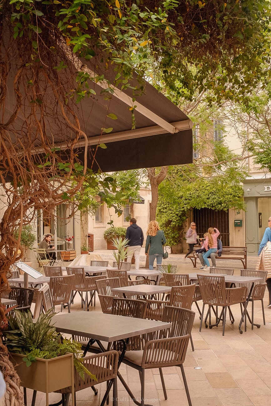 Outdoor space with tables, chairs, and walking people. Vines adorn background buildings, while a large awning stands alongside them in the foreground.