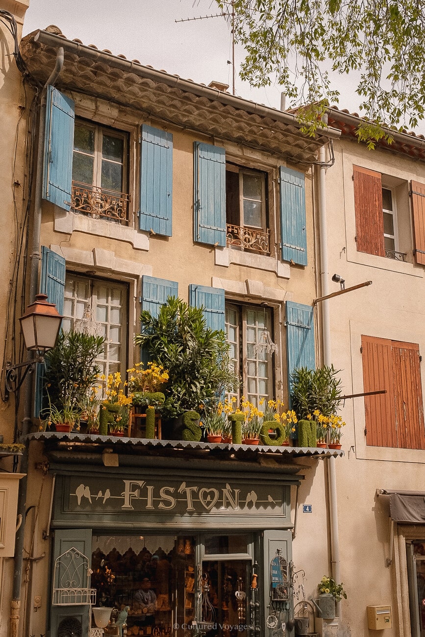 A three-story building in Saint Remy de Provence. The ground floor store has a yellow flower-filled awning and blue wooden shutters on its glass windows.
