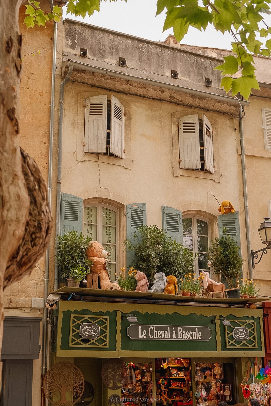 A building with wooden-shuttered windows. The ground floor is adorned with a long green awning featuring rabbit and teddy bear figures, accompanied by potted plants.
