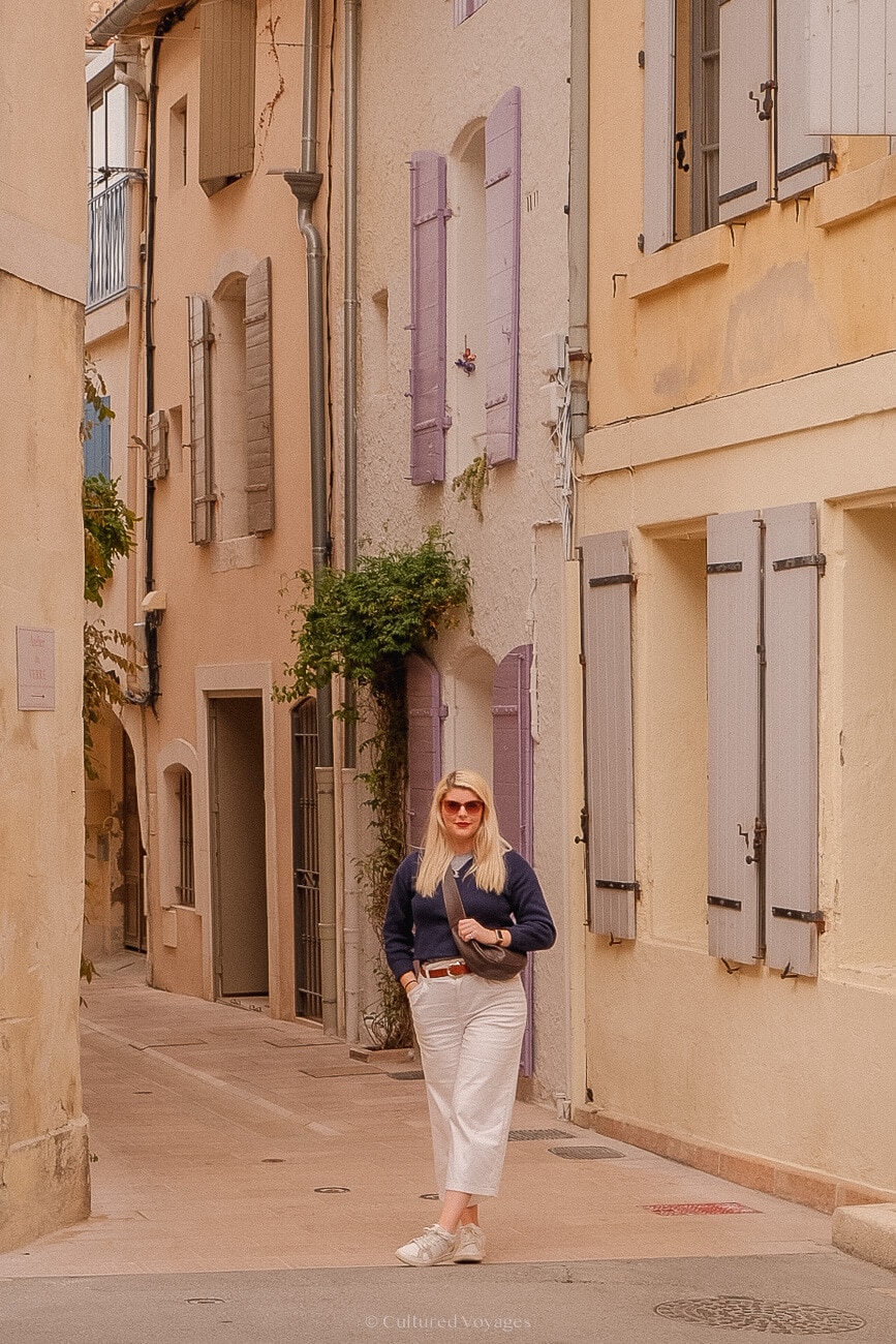 A person standing in the middle of a street lined with warm-colored stone buildings adorned with vines and featuring large windows with wooden shutters.