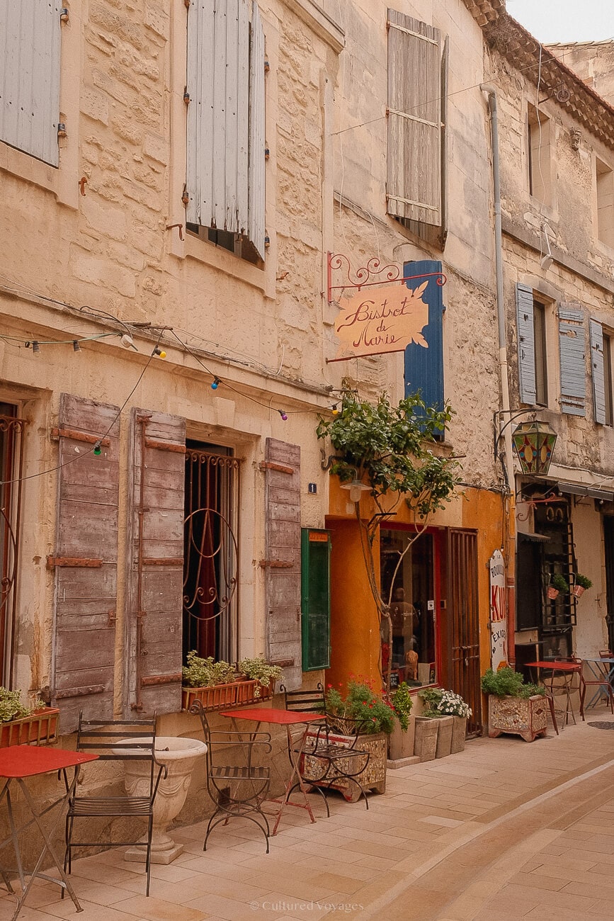 A row of stone bricked buildings in white and yellow, featuring large windows with wooden shutters. Tables and chairs are placed outside along the road you can see when visiting saint remy de provence.