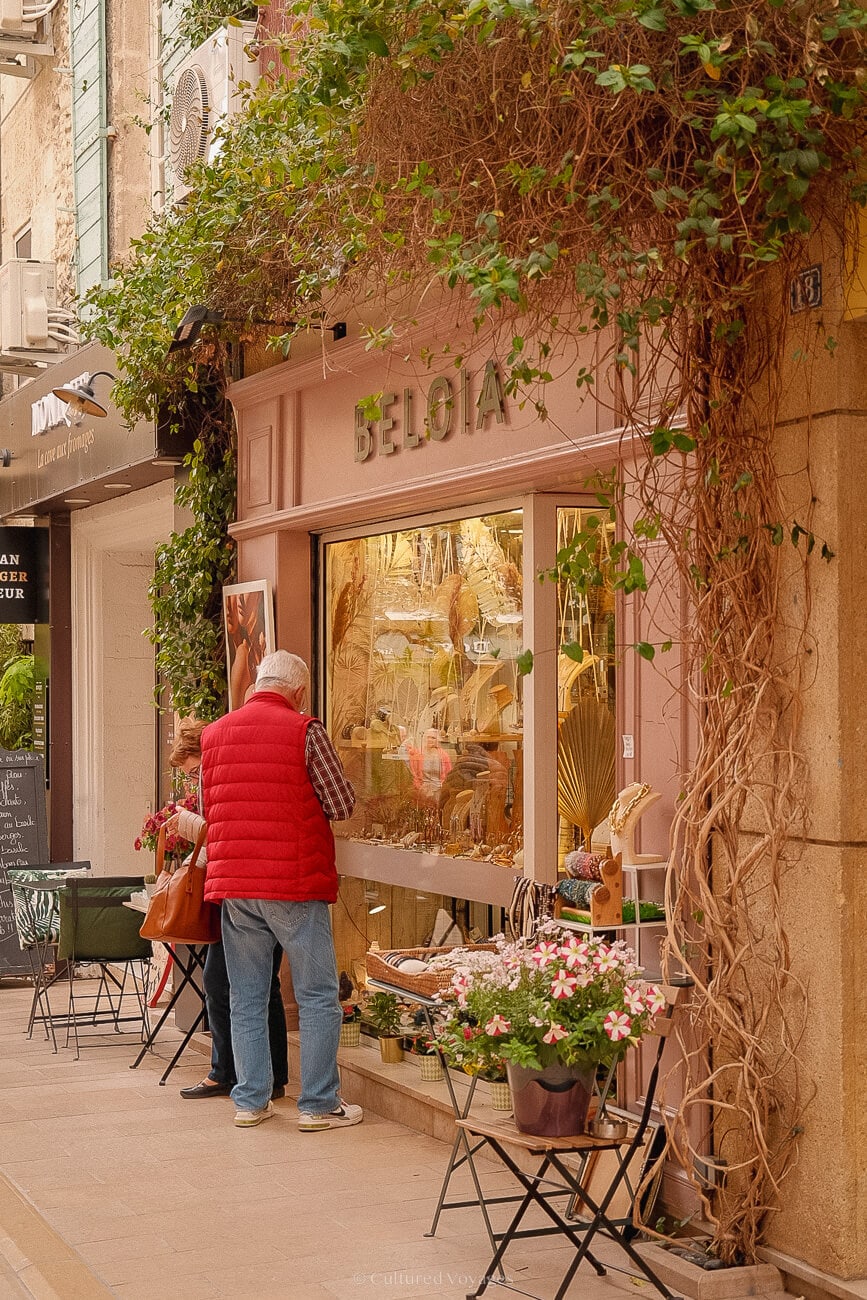 A shop adorned with vines as people peer through its glass windows. Chairs with potted plants and pink flowers line the street.