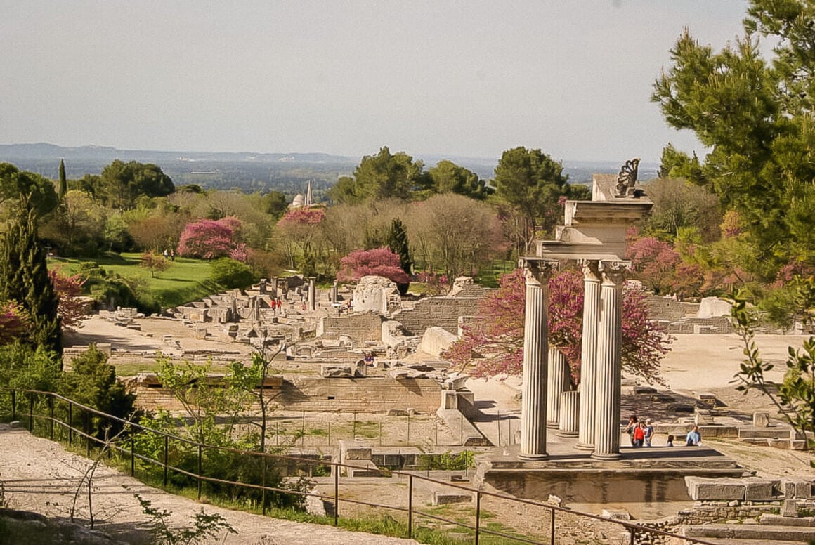 An outdoor exhibition featuring old roman pillars and ruins with people, trees, green fields, pink flowers, and cloudy skies when visiting saint remy de provence.
