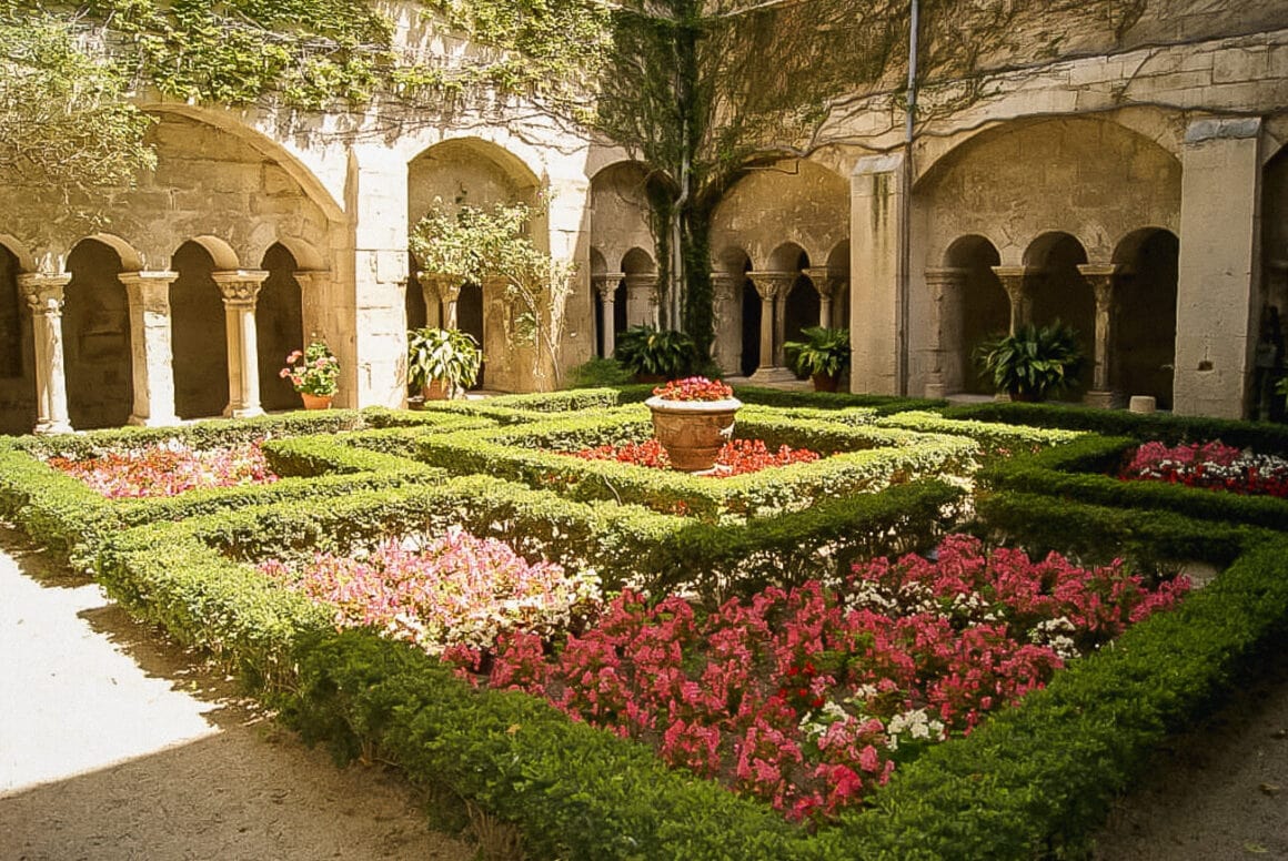 A garden with manicured hedges, vibrant pink and white flowers, potted plants, and walls adorned with arches covered in vines in Saint Rémy de Provence.