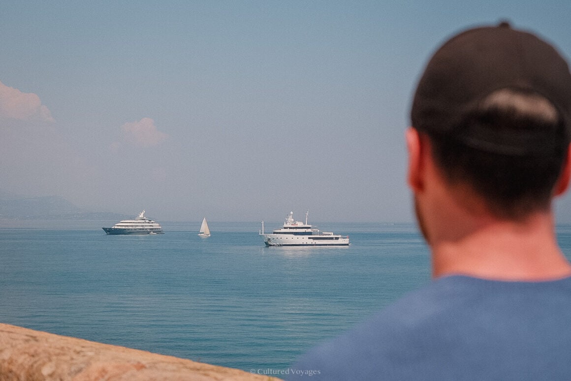 A person in the foreground in a black cap and blue shirt looking at the blue waters in the background with white yachts and a small boat under blue skies.