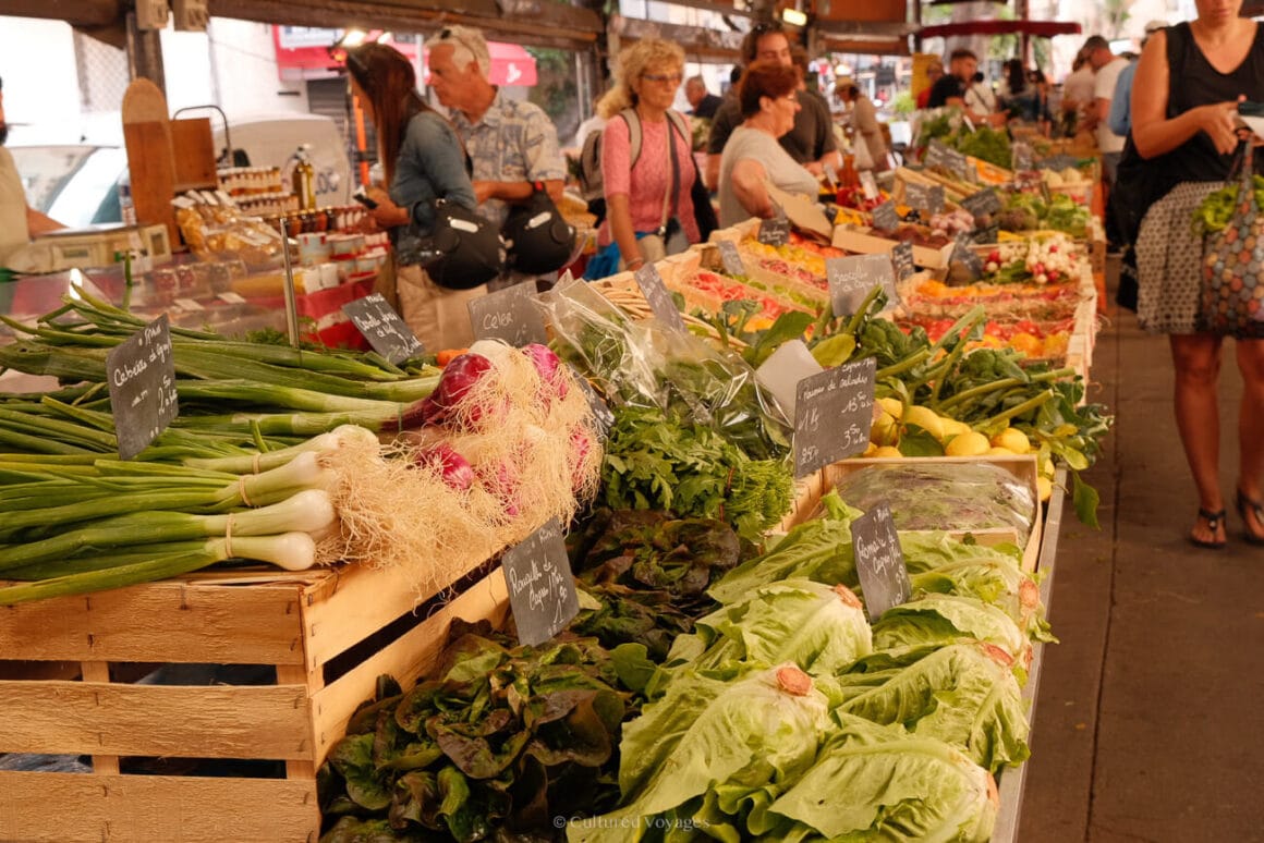 A market filled with displays of vegetables and fruits on wooden crates and people buying, one of the most worth visiting Antibes activities.