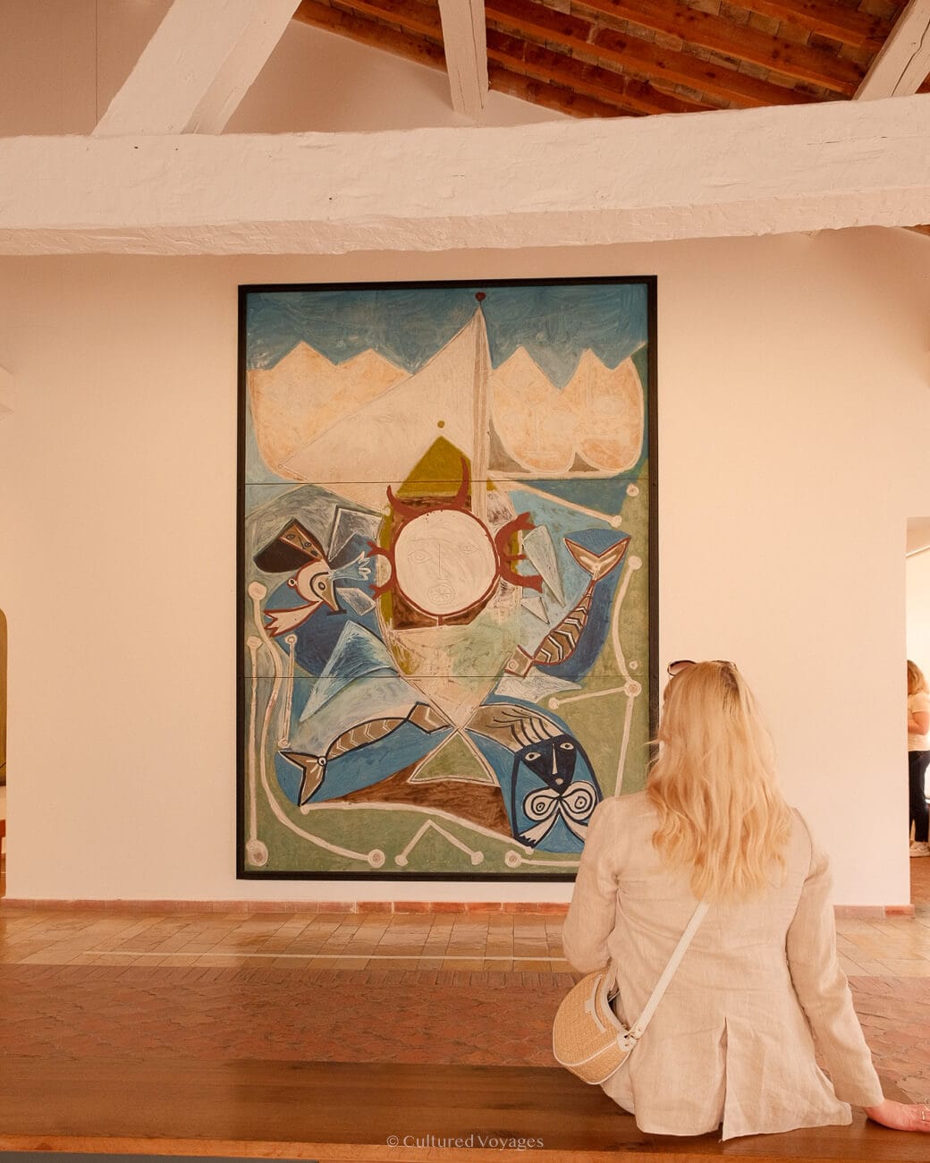 Me wearing a white coat sitting on a wooden bench inside a white walled room with looking at a painting, a worth visiting Antibes activity.