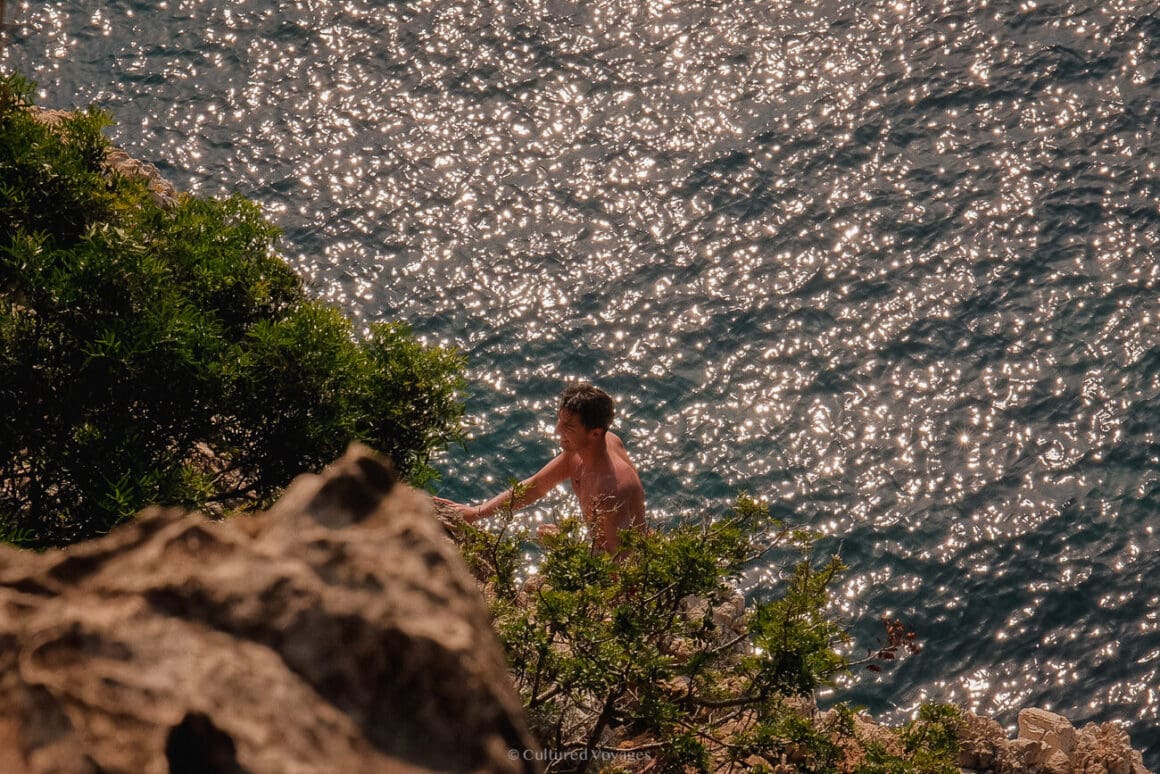 A person climbing the cliff with some small plants and a view of crystal blue waters in the background.