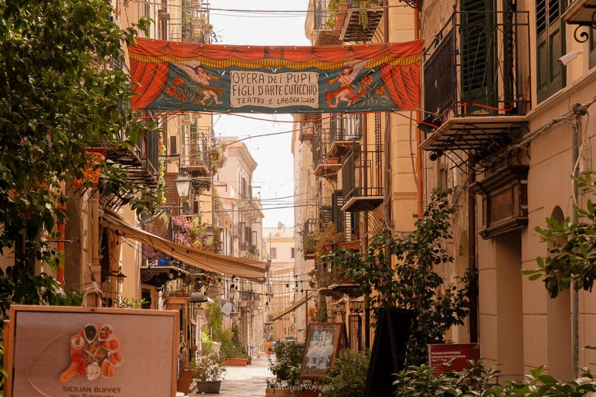 A street filled with potted plants outside the line of buildings with windows and balconies on the upper levels. A banner can also be seen hanging in between two buildings.